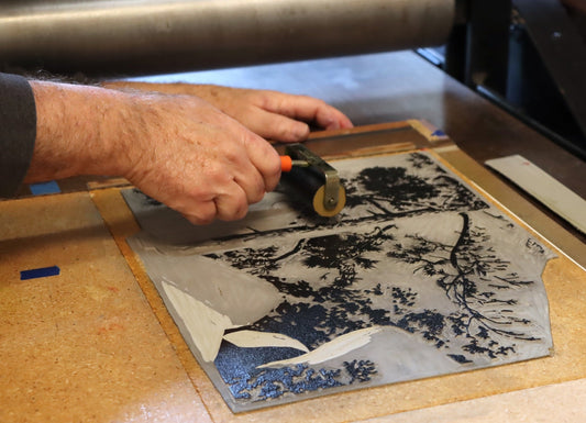  Craig Mitchell inking a Lake Tahoe block print in his Reno studio, rolling black ink across a hand-carved linoleum block showing pine trees and shoreline