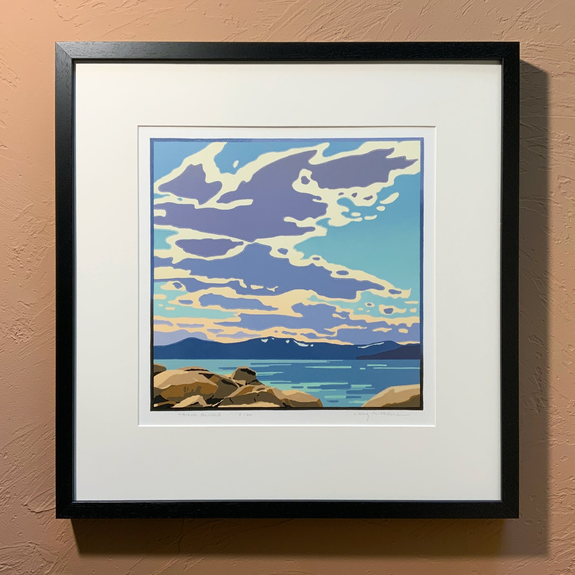 A framed art print depicting a scenic view of Lake Tahoe with clouds in the sky, placed against a neutral wall.