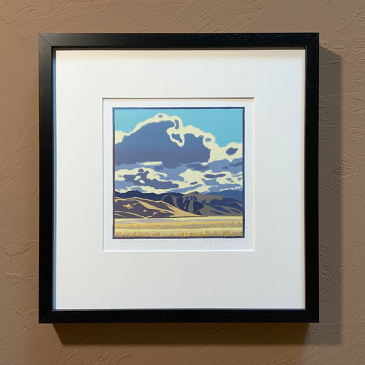 A framed artwork depicting a stylized landscape with clouds in the sky and mountains in the background, primarily in blue and white tones.