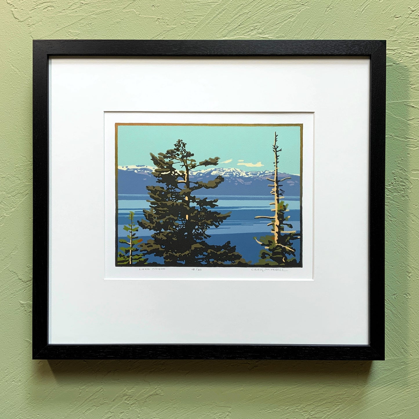 A framed artwork depicting a scenic view of Lake Tahoe with trees and mountains in the background, placed on a green wall.