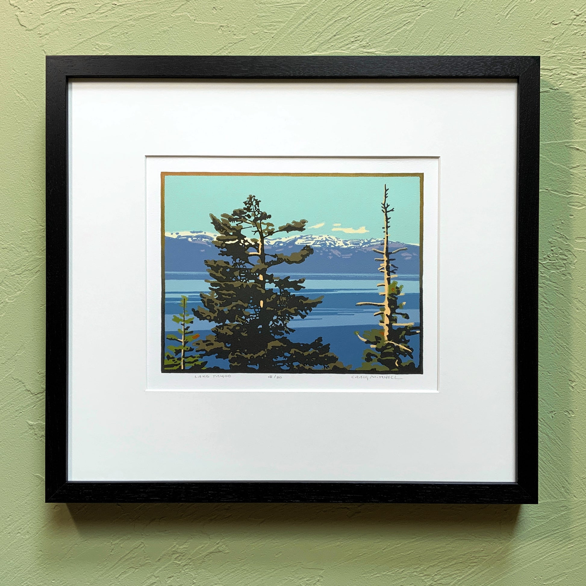 A framed artwork depicting a scenic view of Lake Tahoe with trees and mountains in the background, placed on a green wall.