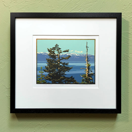 A framed artwork depicting a scenic view of Lake Tahoe with trees and mountains in the background, placed on a green wall.